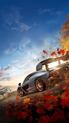 a silver sports car driving down a road surrounded by fall leaves in the background and an overcast sky