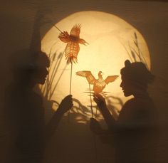 two people holding up paper birds in front of a light that is shining on the wall