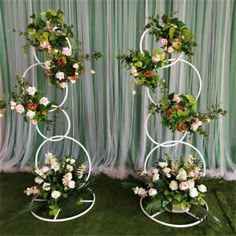 two white metal stands with flowers and greenery on them in front of a curtain