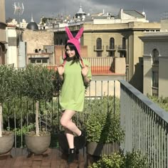 a woman in a green dress and pink bunny ears standing on a balcony with potted plants