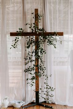 a wooden cross sitting on top of a wooden floor next to a window with sheer curtains