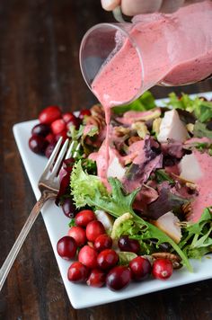 a person pouring dressing onto a salad with cranberries on the side and garnished with pomegranates