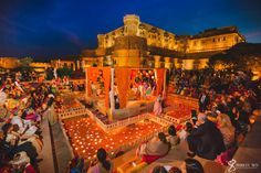 people are sitting around candles in front of a large building at night with lights on the ground