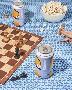 a game of chess and beer on a blue tablecloth next to two cans of soda