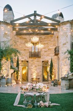 a table with flowers on it in front of a stone building at night, surrounded by string lights