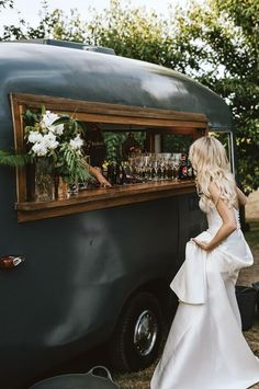 a woman in a white dress standing next to an old fashioned food truck filled with wine glasses