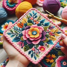 a person holding a crocheted square with flowers on it and balls of yarn in the background