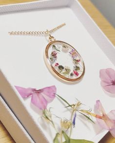 a necklace with flowers on it sitting in a white box next to pink and purple flowers