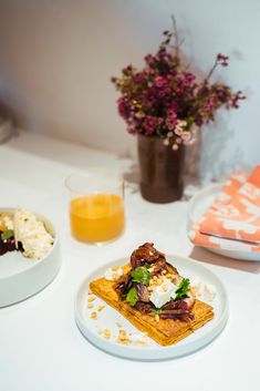 two plates with food on them sitting on a table next to some flowers and orange juice