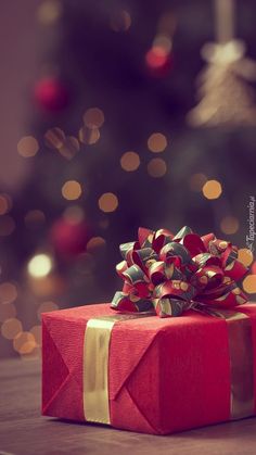 a red present box with gold ribbon and bow sitting on a table in front of a christmas tree