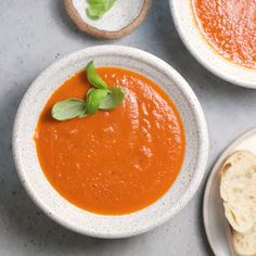 two bowls of tomato soup on a table with bread and basil leaves in the bowl