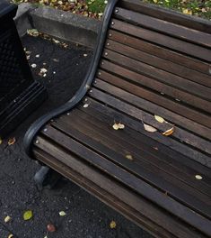 a wooden bench sitting next to a trash can on the side of a road with leaves all over it