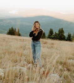 a woman standing on top of a grass covered field next to tall grass and trees