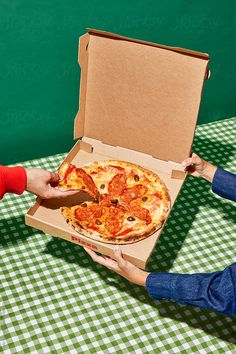 two people holding up a pizza in a box on a table with green and white checkered cloth