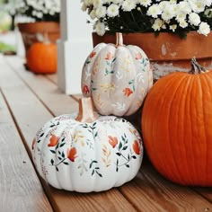 three painted pumpkins sitting on a wooden table next to white flowers and potted plants