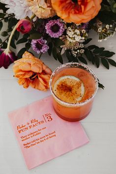 an orange drink sitting on top of a white table next to flowers and a pink napkin