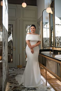 a woman in a white dress standing next to a mirror and vanity with lights on it