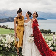 three women standing next to each other in front of some flowers and water with mountains in the background