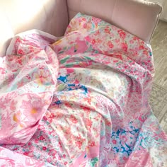 an unmade bed with pink and blue flowers on it's cover, next to a white chair