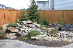 a garden with rocks, plants and water features in the middle of the yard near a wooden fence