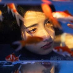 a woman looking at goldfish in an aquarium with her reflection on the water's surface