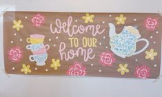 a welcome to our home sign hanging on the wall with flowers and teapots