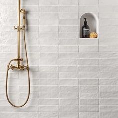 a white tiled bathroom with a gold shower head, soap dispenser and shelf