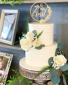 a wedding cake with white flowers and greenery on the table next to a framed photo