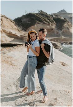 a man and woman standing on top of a mountain looking at a cell phone while holding each other