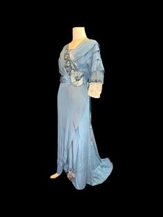 This exquisite antique original Edwardian evening dress, dating back to 1912, features a captivating blue satin fabric that embodies the elegance of its era. The bodice and sleeves are adorned with intricate blue floral patterns complemented by delicate black lace trim, enhancing its visual appeal. Both the front and back necklines showcase an insert of blue chiffon, elegantly finished with a beaded edge, while the neckline and the lower sections of the sleeves are crafted from fine white lace. Elegant Blue Victorian Dress For Evening, Elegant Blue Victorian Evening Dress, Elegant Blue Victorian Floor-length Dress, Blue Elegant Floor-length Victorian Dress, Elegant Blue Floor-length Victorian Dress, Blue Floor-length Victorian Dress, Formal Blue Victorian Dress, Blue Floor-length Victorian Wedding Dress, Victorian Blue Dresses With Historical Design