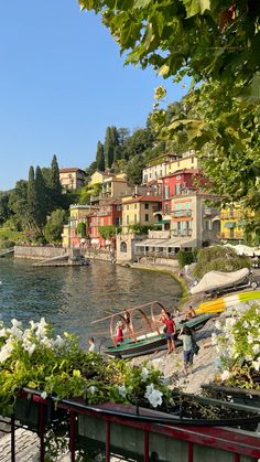 people are walking along the water near some buildings and boats on the shore with flowers in front of them