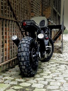 the motorcycle is parked on the cobblestone road by the fence and building behind it