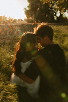 a man and woman are sitting in the grass with their arms around each other as the sun shines behind them