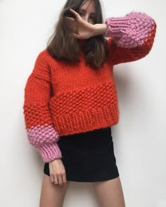 a woman in an orange and pink sweater poses with her hands behind her head while wearing black shorts