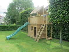 a wooden play house with a slide in the grass next to a green tree and shrubbery