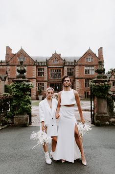 two women standing in front of a large brick building wearing white dresses and holding bouquets