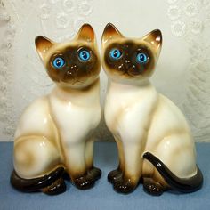 two ceramic cats sitting next to each other on a blue cloth covered table with white lace