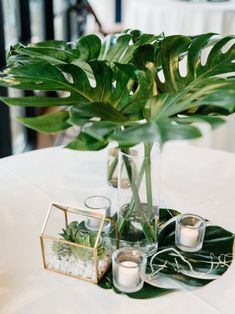 a table topped with a vase filled with green plants and candle holders on top of a white table cloth