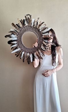 a woman holding a mirror with feathers on it's face and standing in front of a wall