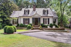 a white house with black shutters in the front yard and trees around it, on a sunny day