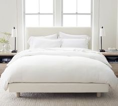 a bed with white linens and pillows in a bedroom next to two large windows