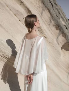 a woman standing in front of a rock formation wearing a white dress with sheer sleeves