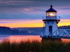 a light house sitting on top of a lake next to tall grass and trees at sunset