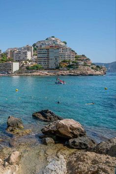 people are swimming in the blue water near some rocks and buildings on top of a hill