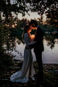 a bride and groom standing next to each other in front of a lake at sunset