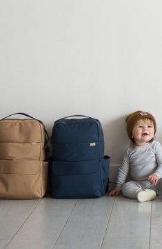 a baby sitting on the floor next to three bags and backpacks, all in different colors