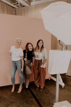 three women standing next to each other in front of a pink wall with an umbrella