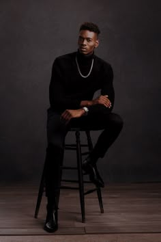 a man sitting on top of a wooden stool wearing a black sweater and pearls necklace