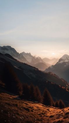 the sun is setting over mountains with trees in the foreground and grass on the ground