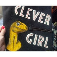 a woman holding up a black bag with a yellow dinosaur on it that says clever girl